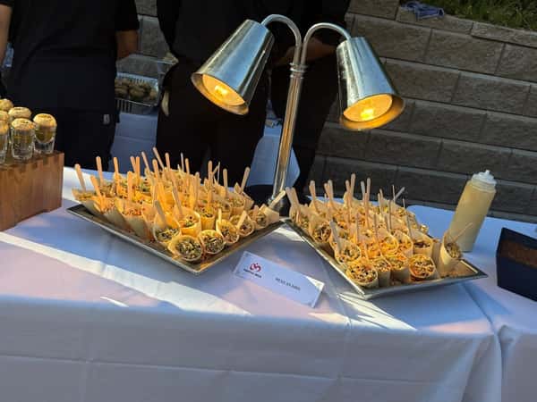 Tray of individual Mexican Bhel servings in paper cones with wooden forks, displayed under heat lamps on a white tablecloth.
