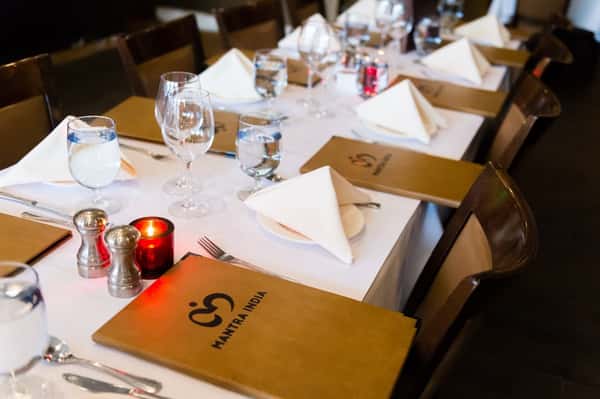 Elegant restaurant table setup with white napkins, wine glasses, water goblets, candles, and Mantra India branded menus