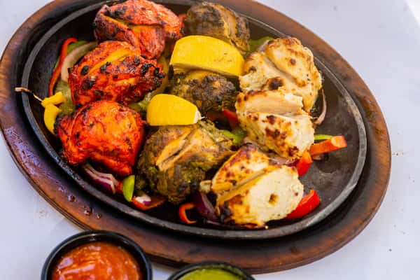 Mixed tandoori chicken platter with red, green, and white marinated pieces served on sautéed peppers and onions with lemon wedges.