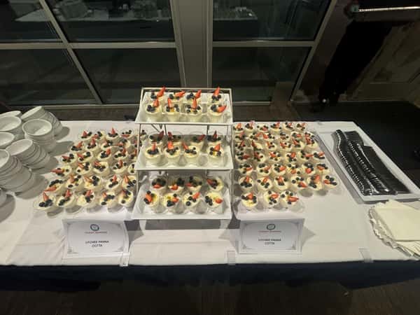 Display of lychee panna cotta desserts topped with berries, arranged neatly on a white tablecloth with bowls, napkins, and cutlery.
