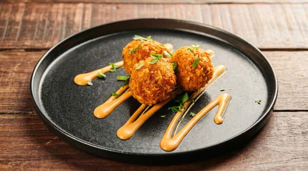 Crispy fried cheese bites served with creamy dipping sauce and fresh herbs on a black plate.