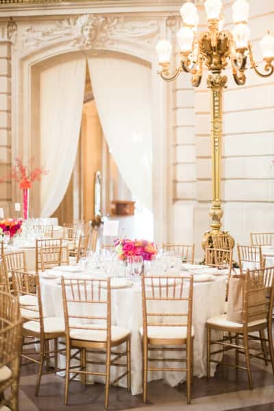 Elegant banquet table with gold chairs, white linens, and vibrant floral centerpiece in a grand hall with ornate architecture.