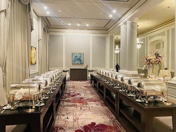 Elegant indoor buffet setup with two rows of chafing dishes on wooden tables, in a ballroom with chandeliers and floral decor.