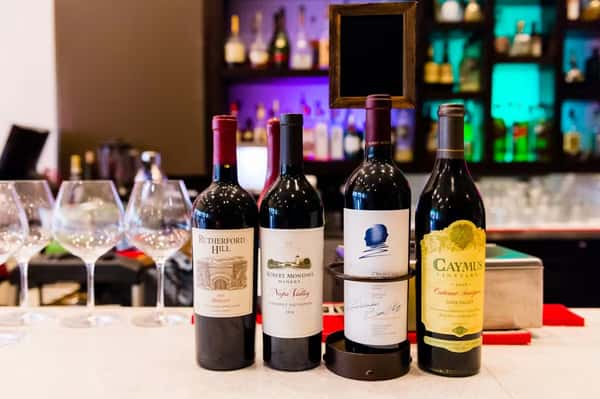 Four bottles of premium red wine with glasses are displayed on a bar counter with colorful backlit shelves.