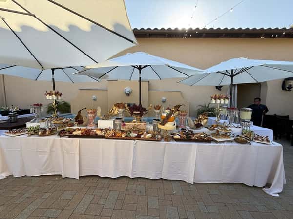 Outdoor buffet setup under large white umbrellas with an elegant display of desserts, snacks, and drinks on white-covered tables.