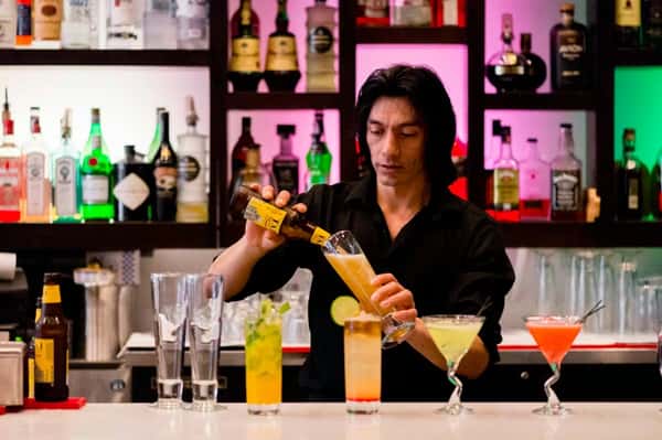 The image shows a bartender skillfully pouring a drink behind a vibrant, well-stocked bar. Several colorful cocktails and glasses are neatly lined up on the counter, and the bar shelves glow with pink, green, and blue lighting, creating a lively and modern ambiance.