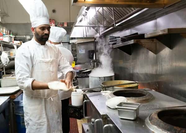 Chef in white uniform prepares naan beside tandoor ovens, with steam rising from pots in a busy commercial kitchen.