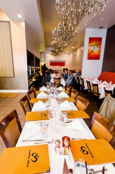 Elegant dining table set with "Mantra India" branded menus, white napkins, and glassware, under ornate chandeliers in a restaurant.