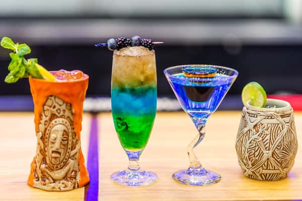 Colorful tropical cocktails in unique glassware, including tiki and carved ceramic mugs, garnished with fruit and herbs.