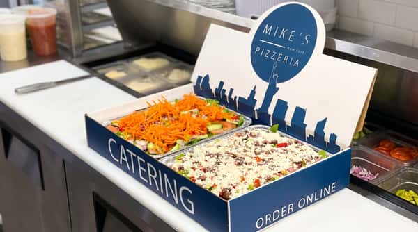 Assorted pizzas and calzones on metal trays, including a round chicken pizza and a square pizza with ricotta and tomato sauce.