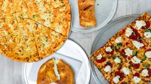 Assorted pizzas and calzones on metal trays, including a round chicken pizza and a square pizza with ricotta and tomato sauce.