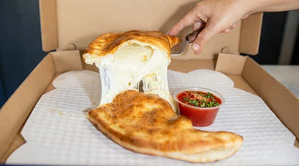 A hand lifting a calzone, revealing stretchy white cheese inside, with a side of red marinara sauce.