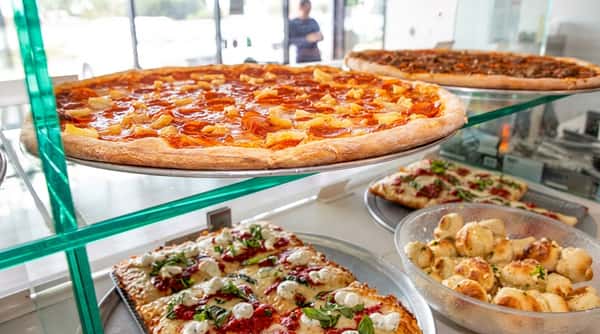 Display case with pepperoni and pineapple pizza, square focaccia slices, and garlic knots.