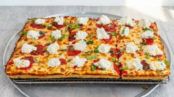 Square-cut Detroit-style pizza with melted mozzarella, dollops of ricotta, tomato sauce, and fresh basil, on a cooling rack.
