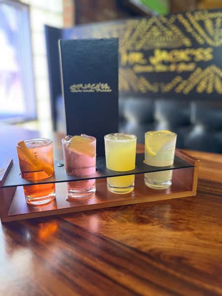 Four cocktails, each a different color and garnish, sit on a tray. The logo "MR. JACK'S" is visible in the background, indicating it's from the bar.