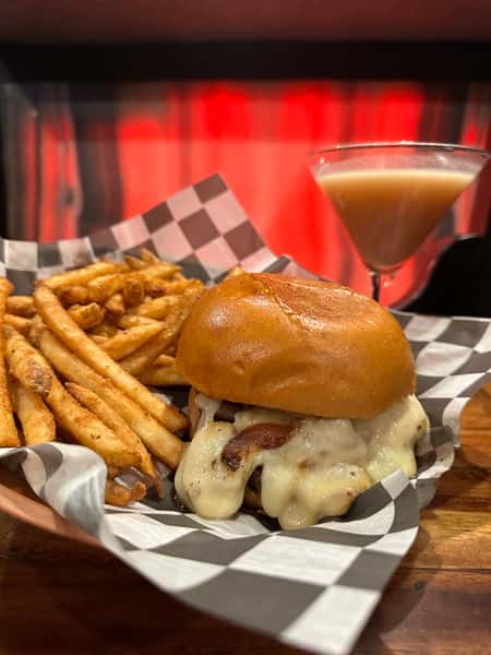 A close up of a cheesy burger and fries with a cocktail behind it, set in a checkered paper-lined dish. This photo comes from the gallery of images of a bar and grill.