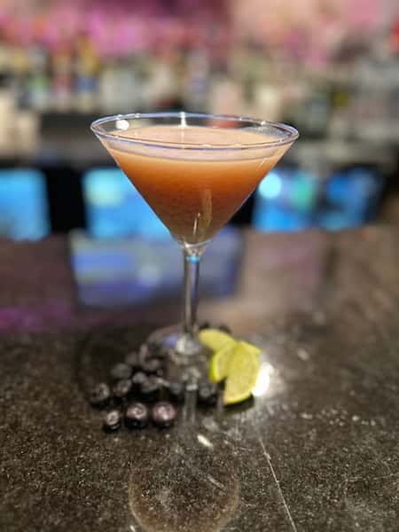 A sophisticated cocktail sits in a martini glass, garnished with fresh blueberries and lime wedges. This image is intended to showcase the drink options at a bar or restaurant.
