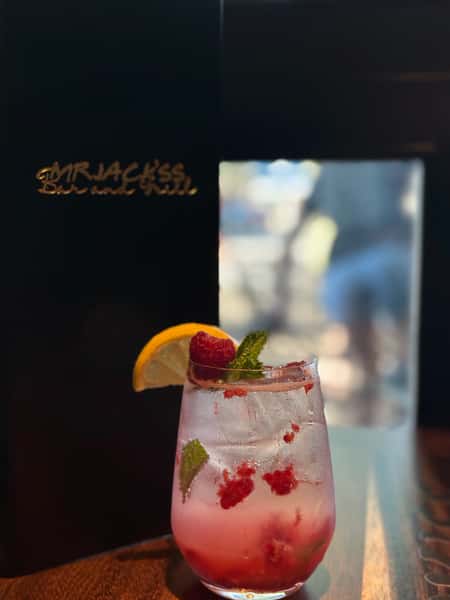 A vibrant cocktail with raspberries and mint is shown in front of Mr. Jacks Sports Bar & Grill menu. The drink is garnished with lemon.