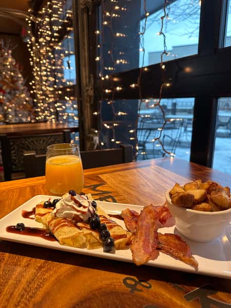 A breakfast meal with blueberry crepes, bacon, and potatoes is presented on a wooden table, alongside a glass of orange juice. String lights and a blurred Christmas tree adorn the background, adding to the restaurant's cozy atmosphere.
