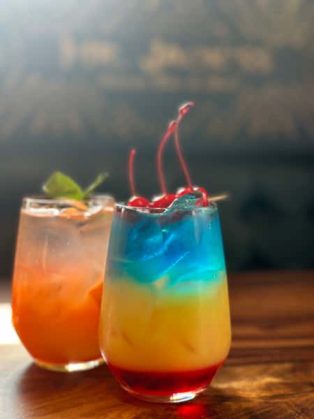 Two cocktails are displayed: one colorful layered drink with cherries, and an orange drink with mint. They're on a wooden surface and a blurry background.