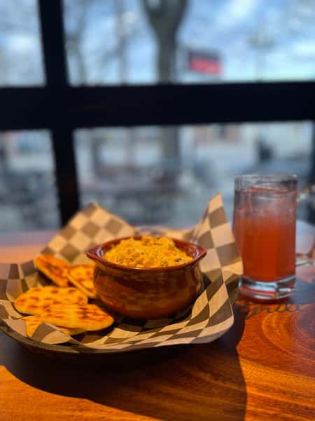 Appetizing image shows a dip served with toasted pita bread alongside a glass of red juice, likely for a restaurant gallery. The dish sits on a checkered napkin on a wood table near a window.