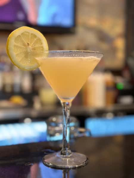 A bright yellow cocktail, possibly a lemon drop martini, in a stemmed glass with a lemon slice garnish. The drink is set on a bar top in a restaurant or bar setting.