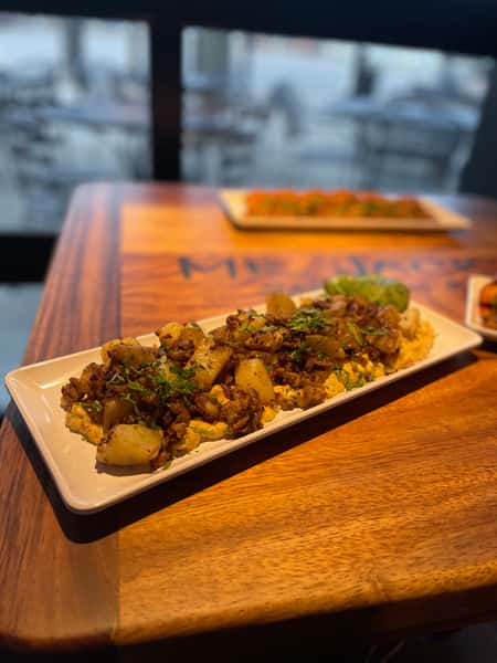 A delectable appetizer features a base of creamy hummus, topped with spiced ground meat, potatoes, and fresh herbs, all served on a sleek white platter. The platter sits on a wooden table, adding warmth to the presentation.