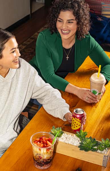 Two women smiling at a table with a smoothie, a can of Poppi soda, and a Good Life Bowls fruit parfait with banana and strawberry.