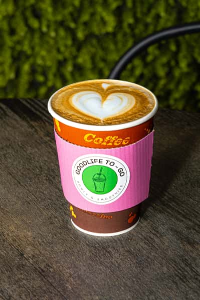 Hot coffee in a to-go cup with latte art and a pink "GoodLife To-Go" sleeve on a wooden table.