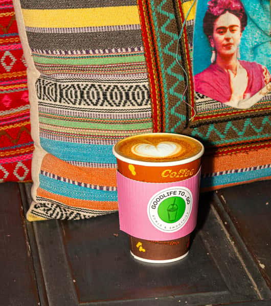 GoodLife To-Go coffee cup with heart-shaped latte art, placed in front of colorful pillows, one featuring Frida Kahlo.