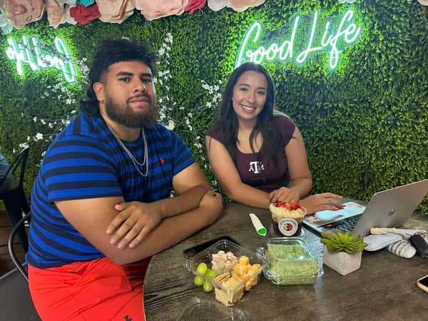 Two people sitting at a table with healthy snacks and a GoodLife Bowl, smiling in front of a neon Good Life sign.