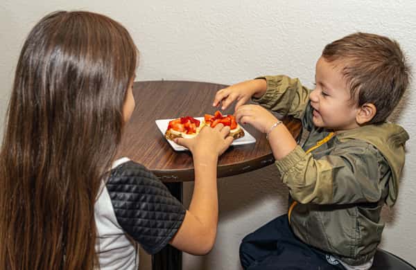 Two young children sitting at a small table, happily sharing toast topped with banana slices and fresh strawberries.