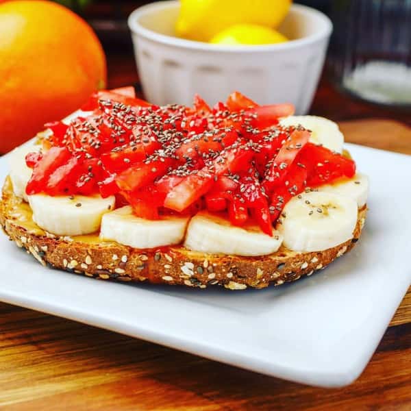 Multigrain toast topped with banana slices, strawberries, and chia seeds on a white plate with citrus fruit in the background.