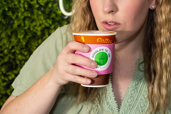 Woman sipping a hot drink from a GoodLife To-Go cup with a pink sleeve and latte art on top.