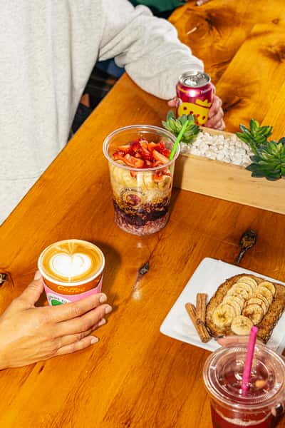 Acai bowl topped with bananas and strawberries, banana toast, and drinks including coffee and strawberry Poppi soda on a wooden table.