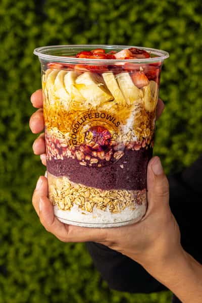 Layered smoothie bowl in a clear cup with granola, banana, strawberries, and acai, held in front of a green leafy backdrop.