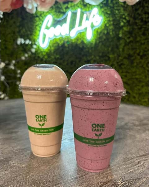 Two One Earth smoothies, one pink and one beige, sit on a table in front of a neon Good Life sign and a green floral wall.