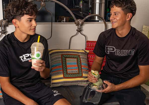 Two teenage boys in athletic gear sitting on a bench, one drinking a smoothie and the other holding a fresh wrap with a smile.