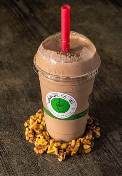 Chocolate smoothie from GoodLife To-Go with a red straw, surrounded by walnuts on a rustic wooden surface.