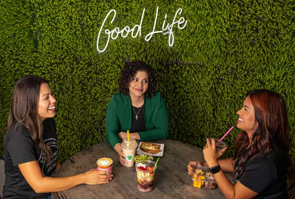 Three women sit at a round table under a "Good Life" sign, enjoying smoothies, coffee, toast, and fruit snack packs.