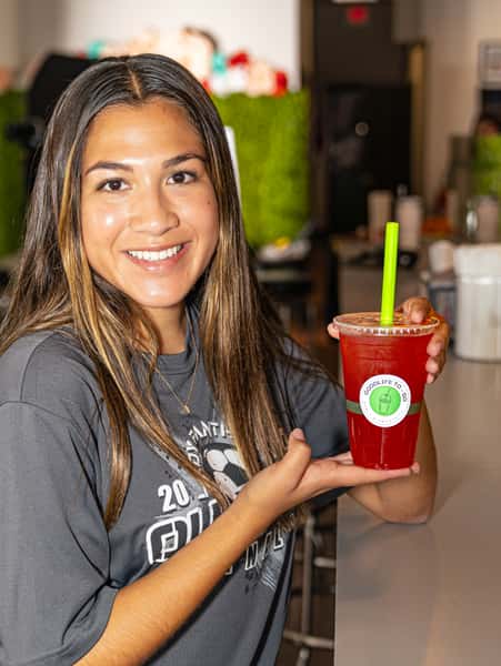 Smiling woman holding a red iced drink with a green straw from GoodLife To-Go inside a bright, modern cafe.