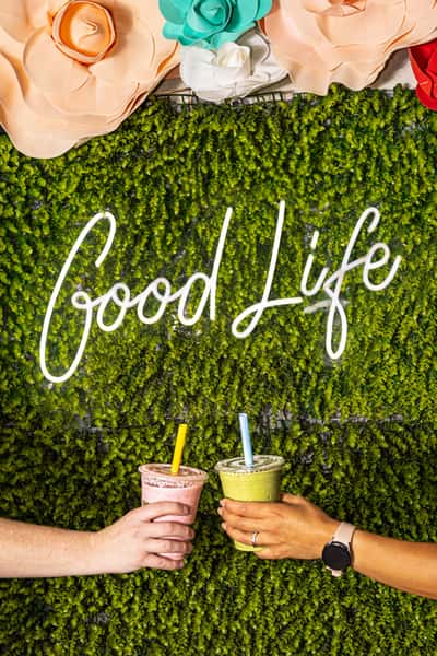 Two people holding smoothies in front of a green wall with a neon "Good Life" sign and decorative flowers.