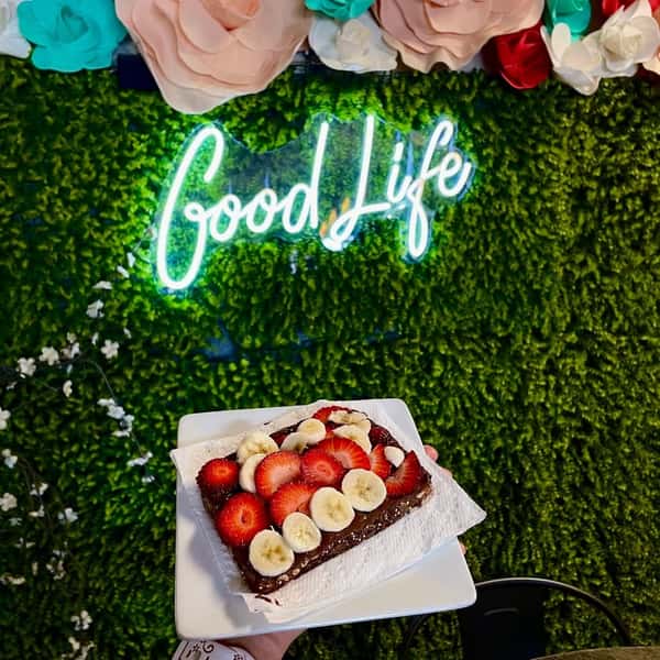 Chocolate toast topped with banana and strawberry slices held in front of a neon Good Life sign and floral green wall.