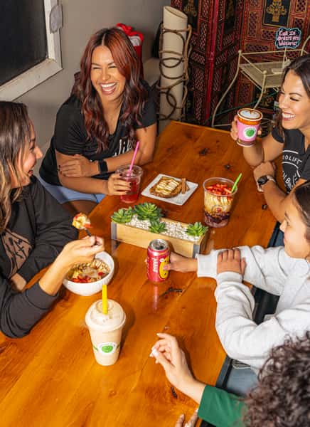 Group of friends enjoying smoothies, acai bowls, toast, and drinks around a wooden table with bright smiles and lively conversation.