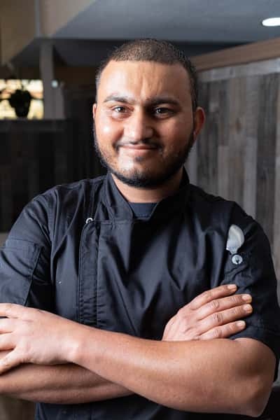 A chef stands smiling confidently with arms crossed, wearing a black chef coat with a fork emblem on the sleeve.
