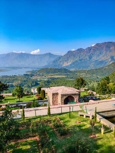 This image features a beautiful terraced Mughal-style garden with manicured lawns, flower beds, and a stone pavilion, set against a stunning backdrop of the Himalayas and a lake below, under a bright blue sky.