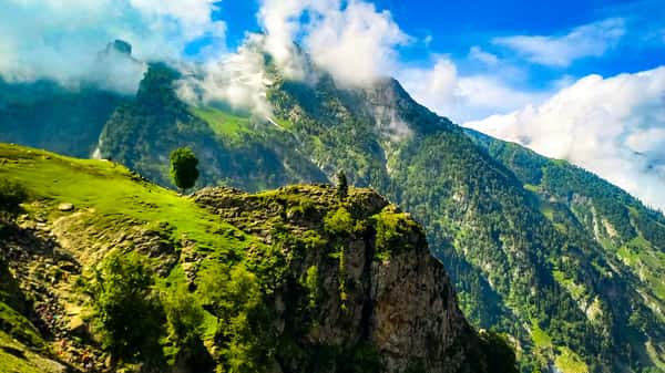 Lush green mountains with scattered trees, rocky cliffs, and clouds drifting around snowy peaks under a vibrant blue sky.