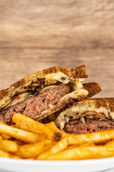 Close-up of a juicy patty melt sandwich stuffed with grilled mushrooms, melted cheese, and beef on crispy toasted rye bread, served with golden French fries on a white plate.