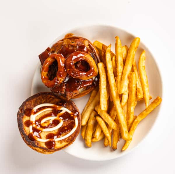Overhead view of a BBQ bacon cheeseburger topped with crispy onion rings and drizzled with barbecue sauce, served open-faced alongside a generous portion of golden French fries on a white plate.