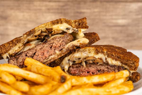 Close-up of a juicy patty melt sandwich stuffed with grilled mushrooms, melted cheese, and beef on crispy toasted rye bread, served with golden French fries on a white plate. Ask ChatGPT